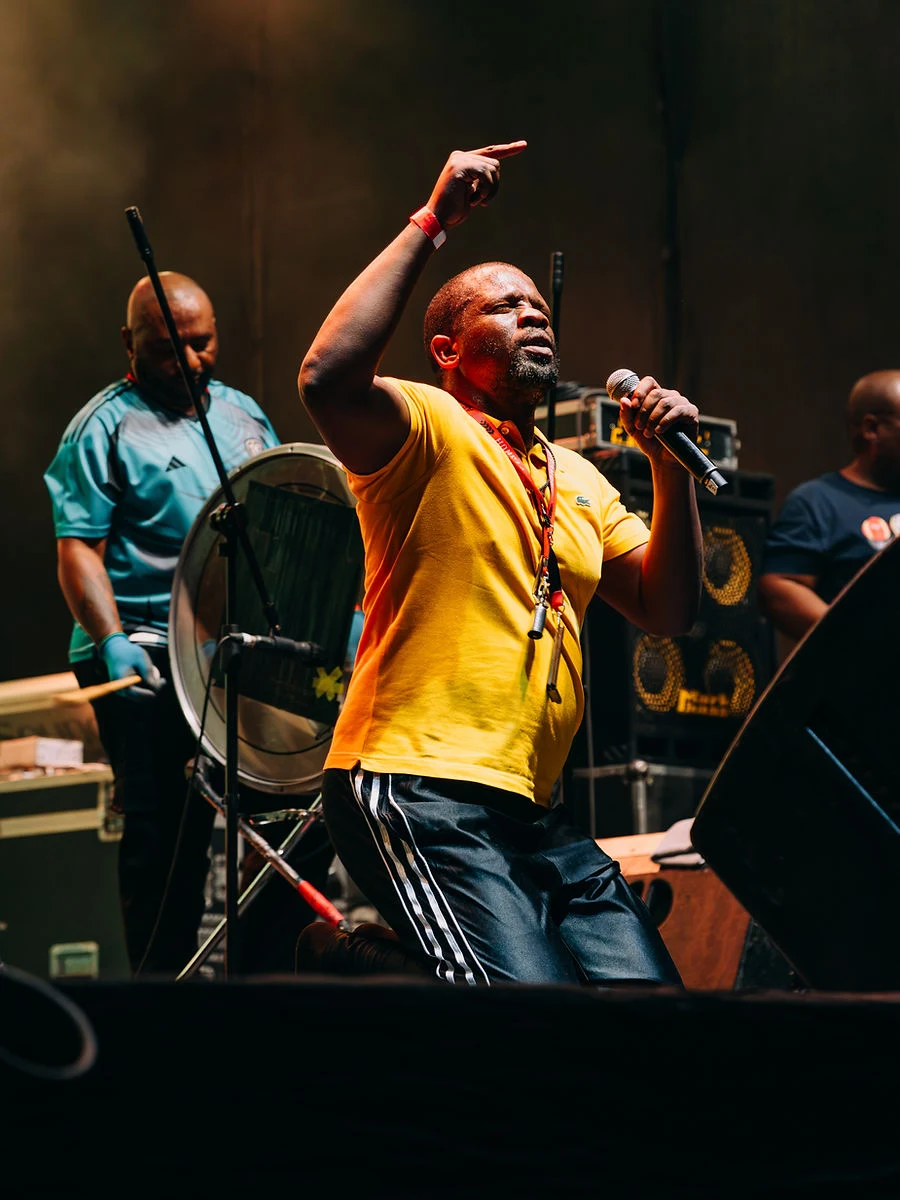 A performer in a yellow shirt kneeling on stage, finger pointed to the sky, microphone in hand